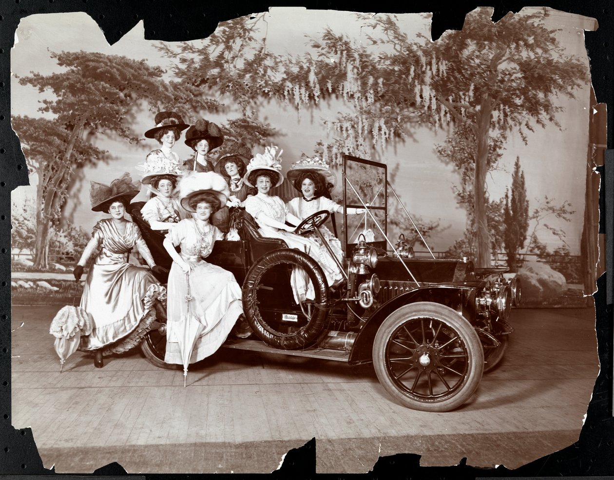 Kahdeksan pukeutunutta naista poseerasi autossa ja sen ympärillä näyttämöllä maisema taustalla, oletettavasti esityksessä "The Vanderbilt Cup", 1906 (hopea gelatiiniprintti) tekijältä Byron Company