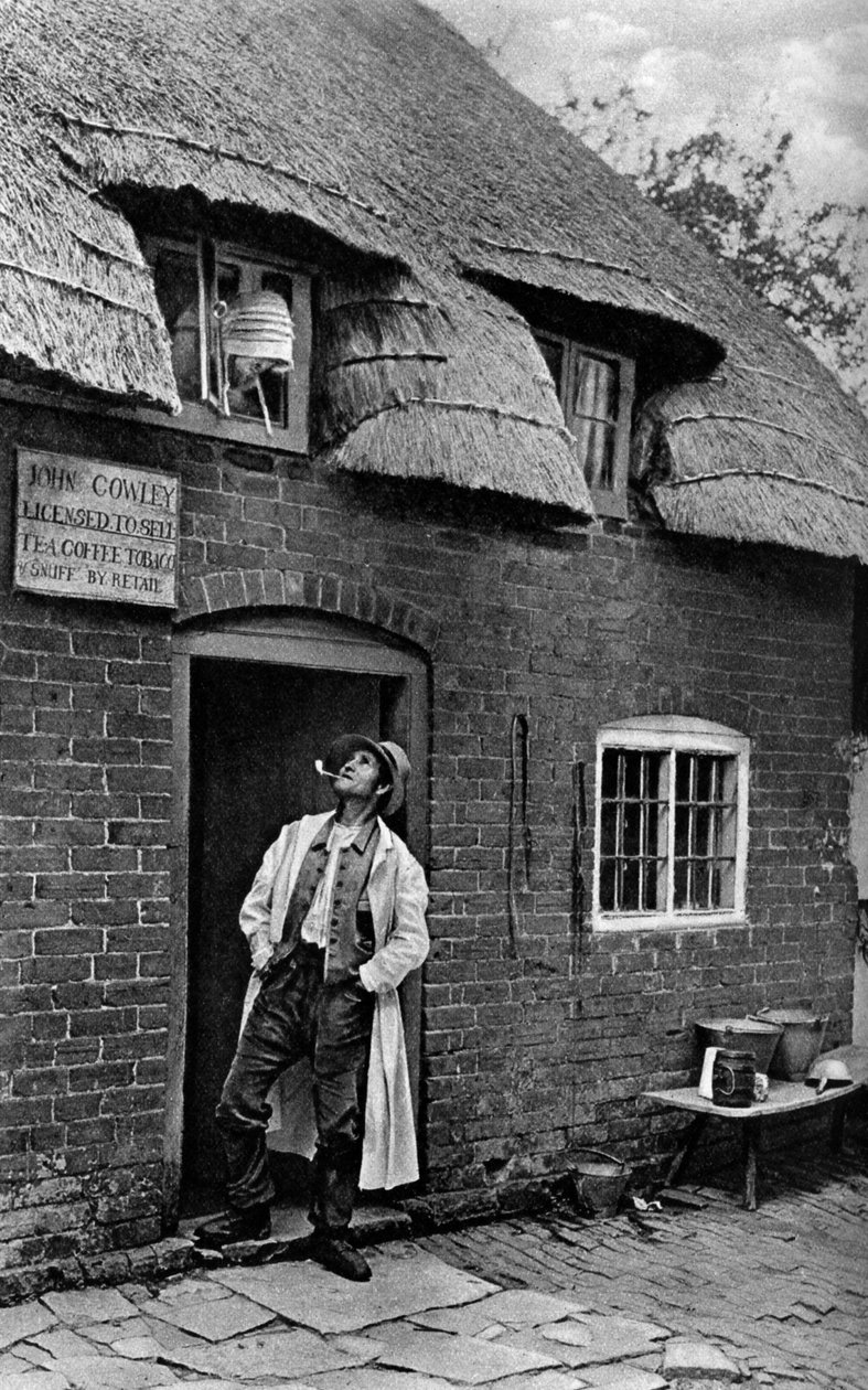 Mies polttaa piippua liikkeen ulkopuolella, Worcestershire, vuodelta 1922. tekijältä A. W. Cutler