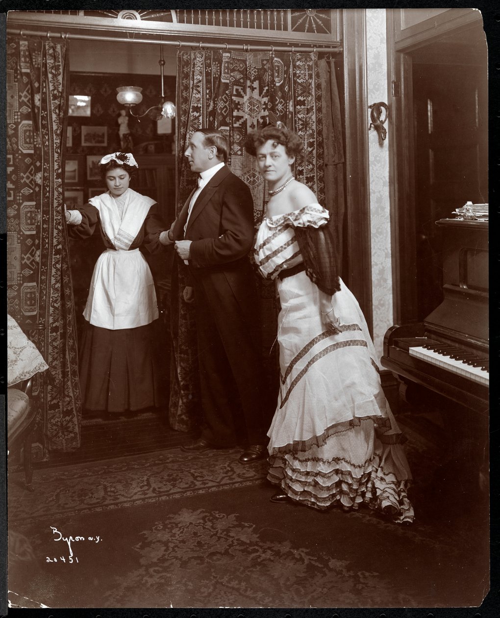 Muodollisesti pukeutunut mies ja nainen sekä kotipalvelija huoneessa, jossa on piano, 1905 (hopeagelatiiniprintti) tekijältä Byron Company