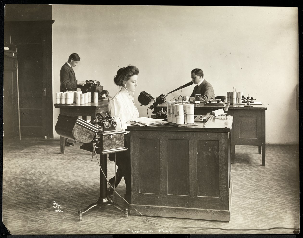 Sisäkuva naisesta, joka kirjoittaa sanelukoneella, ja miehestä, joka sanelee toiselle Edison Phonograph Co.:ssa, West Orange, New Jersey, 1903 (hopeagelatiiniprintti) tekijältä Byron Company
