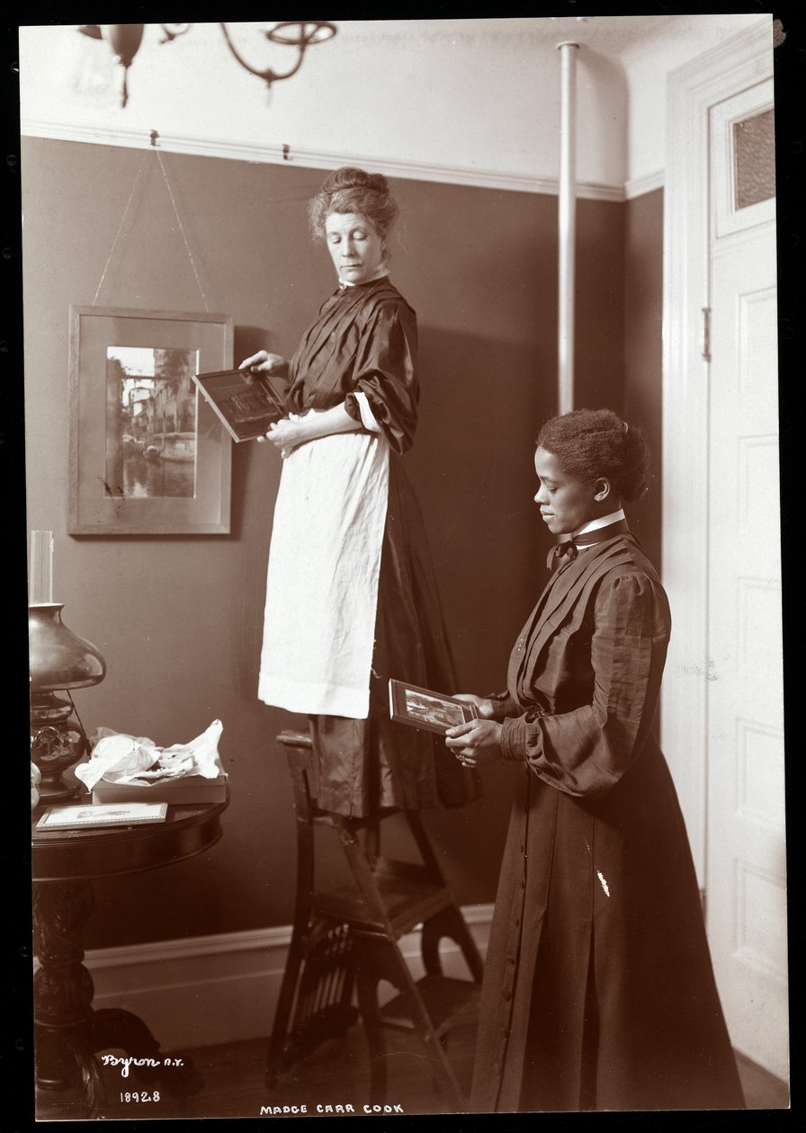 Madge Carr Cook ripustaa kuvia piikansa kanssa, n. 1904 (hopeagelatiiniprintti) tekijältä Byron Company