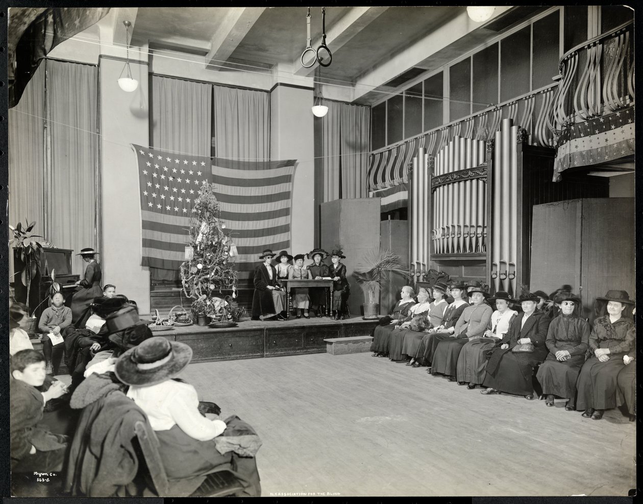 Ihmisiä New Yorkin sokeiden yhdistyksen auditoriossa joulukuusen kanssa ja ihmiset, jotka mahdollisesti esittävät sketsin lavalla urkujen kanssa, 1920 (hopeagelatiiniprintti) tekijältä Byron Company