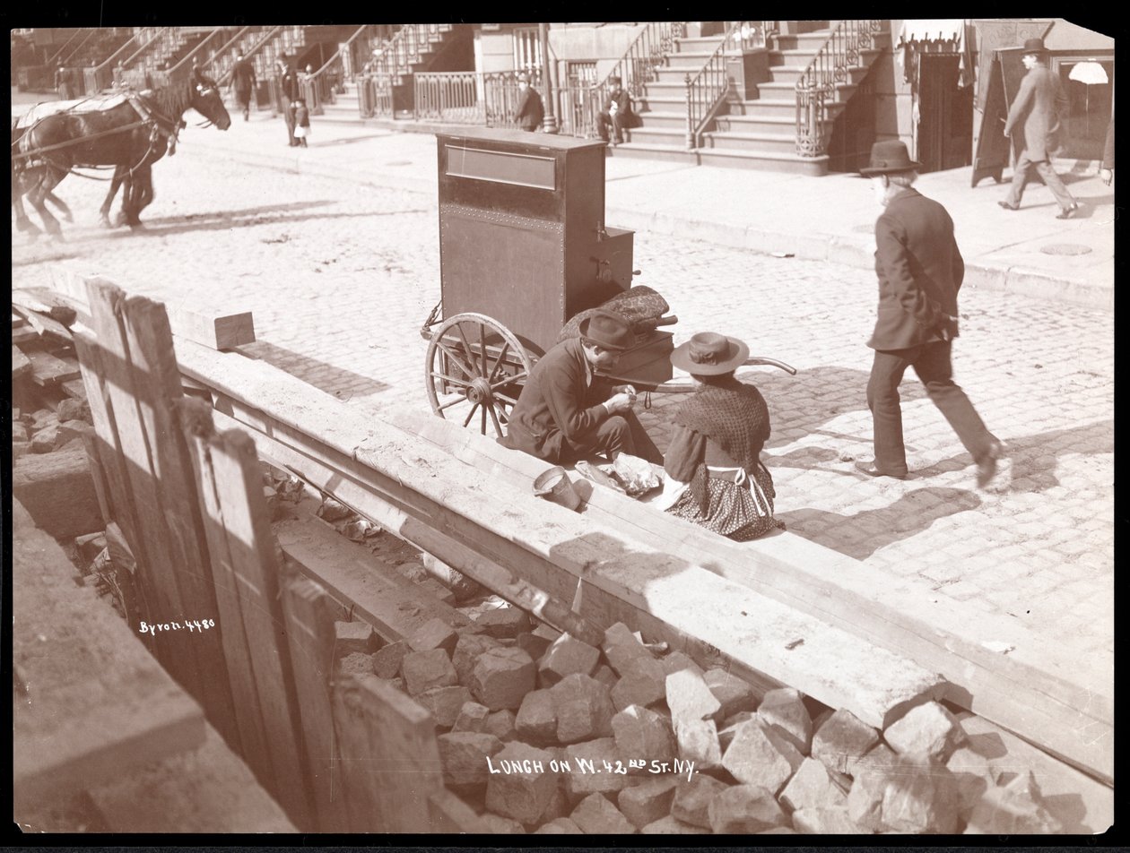 Näkymä katusoittajasta ja naisesta syömässä lounasta West 42nd Streetillä, New York, 1898 (hopeagelatiinivedos) tekijältä Byron Company