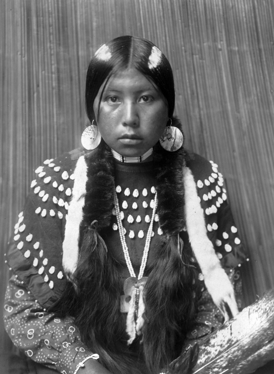 Kalispel-intiaaninainen, hiuksiin maalatut valkoiset raidat, kuorilevykorvakorut, yllään hirvenhampailla koristeltu huopamekko, n. 1910, valokuva Edward S. Curtis tekijältä Edward Sheriff Curtis