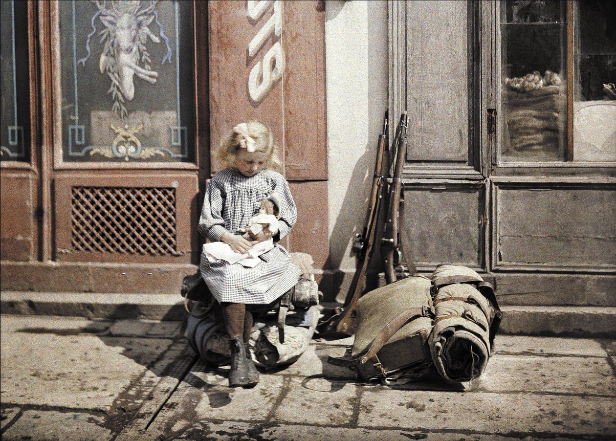 Pieni tyttö leikkii nukkensa kanssa; kaksi asetta ja reppu ovat hänen vieressään maassa, Reims, Marne, Ranska, 1917 (autochrome) tekijältä Fernand Cuville