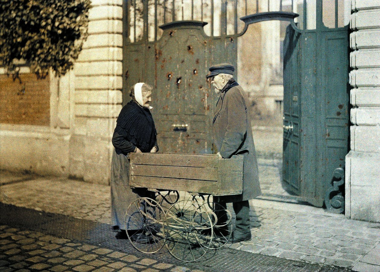 Kaksi Reimsin vanhusta, vanha nainen ja herra Reiser, seisovat portin edessä, jossa on paljon luodinreikiä, Reims, Marne, Ranska, 1917 (autokromi). tekijältä Fernand Cuville