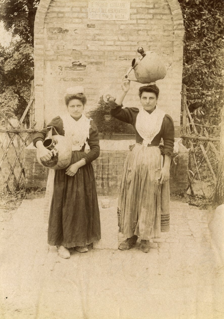 Kaksi nuorta naista perinteisiin vaatteisiin pukeutuneena ja terrakottapurkkia kantaen kaivon lähellä, Marche, Italia, n. 1910. tekijältä Italian Photographer