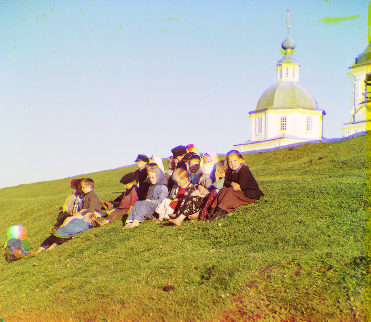 Venäläiset lapset istuvat kukkulan puolella lähellä kirkkoa ja kellotornia maaseudulla lähellä White Lakea, Venäjän pohjoisosassa, 1905-1915 tekijältä Sergey Prokudin Gorsky