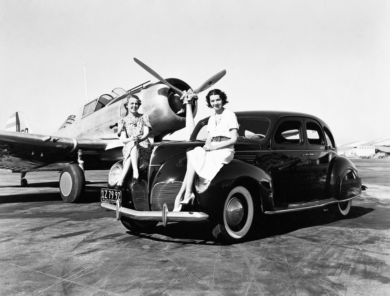 Kalifornia: 1939 Kaksi naista istuvat vuoden 1939 Lincoln Zephyr -sedanin lokasuojissa lentokentällä, ja heidän takanaan on pysäköity yksimoottorinen kone (m/v kuva) tekijältä Unbekannt