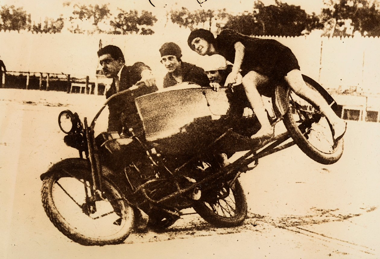 Mies esittelee moottoripyörällä ajamista, jonka sivuvaunussa on kolme nuorta naista. Sivuvaunu nousee maasta, kun he kääntyvät. tekijältä Unknown photographer