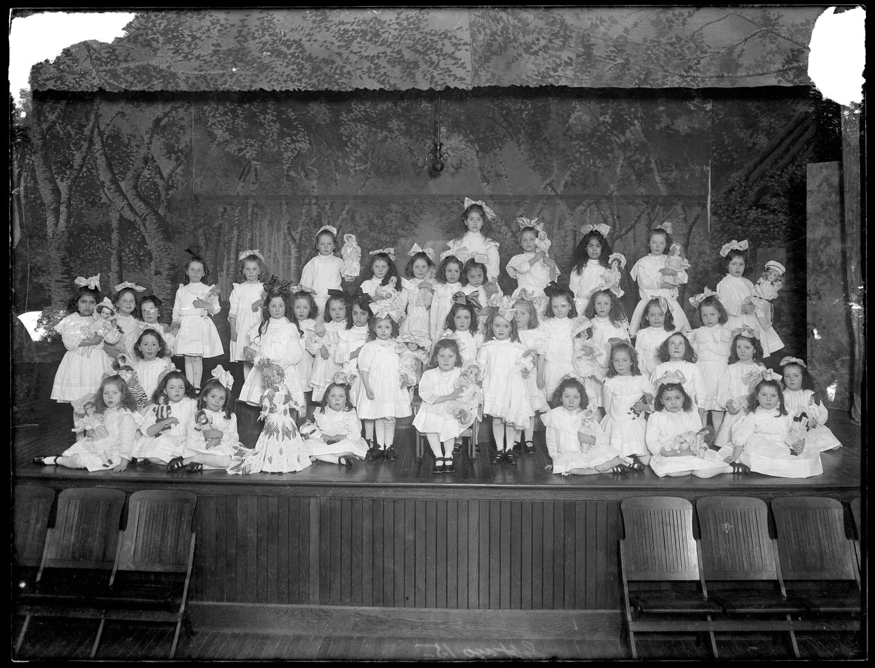 Ryhmä pieniä tyttöjä poseerasi pienellä näyttämöllä pitäen nukkeja, luultavasti roomalaiskatolinen orpojen turvapaikka, Kingsbridge, Bronx, n. 1911 tekijältä William Davis Hassler