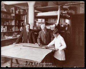 Muotokuva Henrystä ja S.G. Lindemanista poikansa tai pojanpoikansa kanssa seisomassa myymäläpiirustusten päällä pianolle, 1910 (hopeagelatiiniprintti)