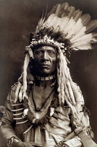 Rabo de Doninha (Apohsuyis). Retrato de cacique da tribo Piegan usa o famoso cocar de penas de águia e de doninha, túnica de camurça, dois colares, um de osso e outro de garras de urso, e uma machadinha de cachimbo tekijältä Edward Sheriff Curtis