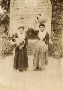 Kaksi nuorta naista perinteisiin vaatteisiin pukeutuneena ja terrakottapurkkia kantaen kaivon lähellä, Marche, Italia, n. 1910. tekijältä Italian Photographer
