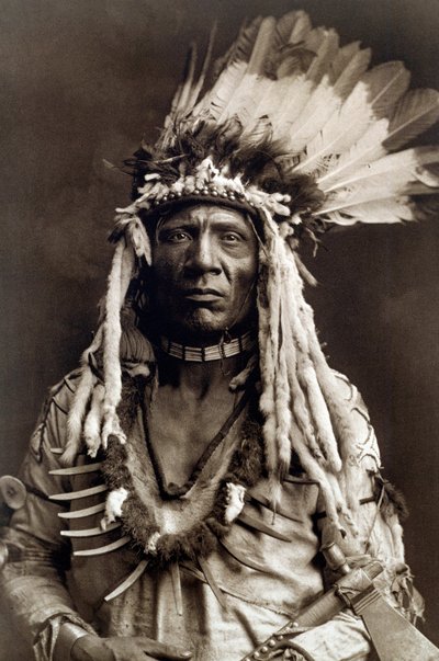 Rabo de Doninha (Apohsuyis). Retrato de cacique da tribo Piegan usa o famoso cocar de penas de águia e de doninha, túnica de camurça, dois colares, um de osso e outro de garras de urso, e uma machadinha de cachimbo tekijältä Edward Sheriff Curtis