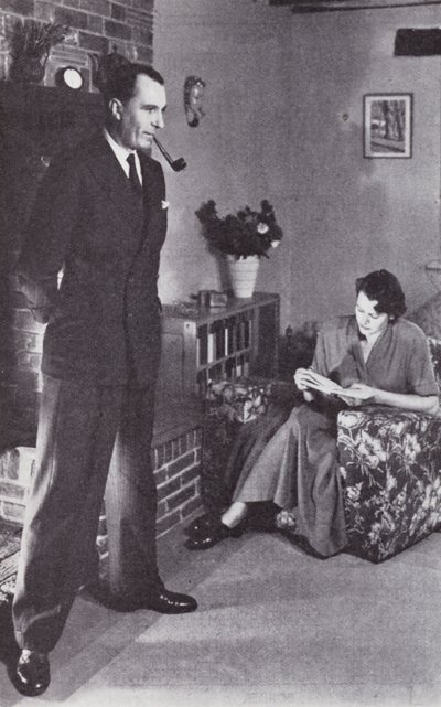 Vaimo lukee ja mies lämmittää selkänsä tulta vasten, n. 1950 tekijältä English Photographer