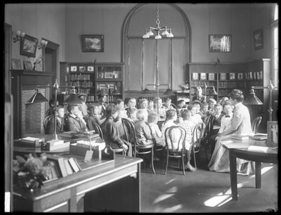 Lapset kuuntelevat naista Queens Borough Public Libraryn tunnistamattoman haaran lukusalissa, n. 1910 tekijältä William Davis Hassler