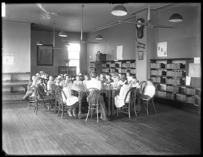 Lapset kuuntelevat naista Queens Borough Public Libraryn tunnistamattoman haaran lukusalissa, n. 1910 (m/v kuva) tekijältä William Davis Hassler
