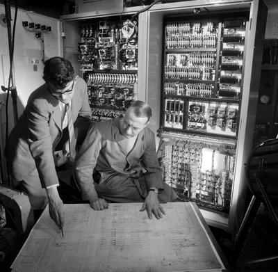 Insinöörit keskustelevat suunnitelmista rakentamansa elektronisen ohjauspaneelin edessä tekijältä Frederick (attr. to) Barnard