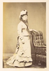 Japanilainen nainen eurooppalaisessa mekossa, Täyspitkä seisova studiomuotokuva nuoresta japanilaisesta naisesta länsimaisessa mekossa ja hatussa,  Valokuvaaja: Tuntematon  Lucas Collection: Valokuvaennätys Charles Jamesin ja Morton P, Lucasin maailmankier