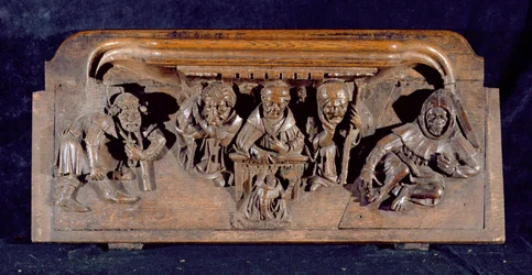Kurjuus, joka kuvaa akateemikkoa istumassa paneelipöydän ääressä ja pitämässä varoitusta polvistuvalle katuvalle, New College Chapel, Oxford, 1300-luvun loppu (puu)