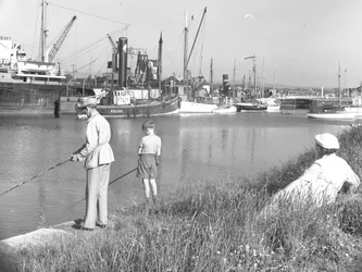 Mies ja poika kalastavat Glasson Dockissa, kun heidän takanaan istuva nainen katselee, erilaisia veneitä on ankkuroituna