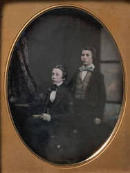Kaksi nuorta miestä rusetissa, yksi istuu kirjaa kädessään, toinen seisomassa, 1850-luku.