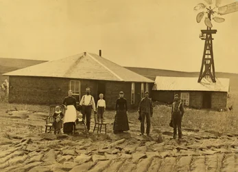 Uudisasukkaiden perhe turvemajansa edustalla, viereisen rakennuksen katolla tuulimylly, Custer County, Nebraska, noin 1885