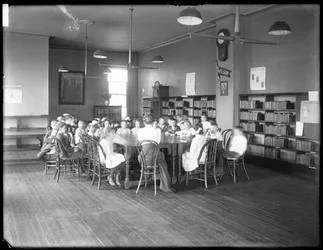 Lapset kuuntelevat naista Queens Borough Public Libraryn tunnistamattoman sivukirjaston lukusalissa, noin 1910