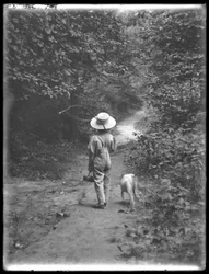 William Gray Hassler olkihatussa ja haalareissa, onkivapa ja kori kantamassa, kävelemässä polkua metsässä pienen koiran kanssa (Bessie), 20. elokuuta 1911